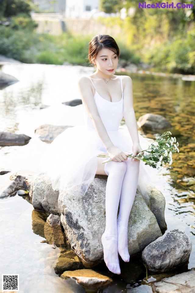 A woman in a white dress sitting on a rock by the water.